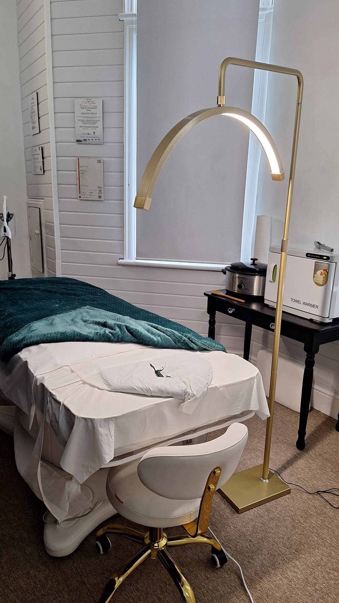 The Beauty Bank Clinic treatment room — gold arc lamp, white panelled walls, branded pillow on the treatment bed.