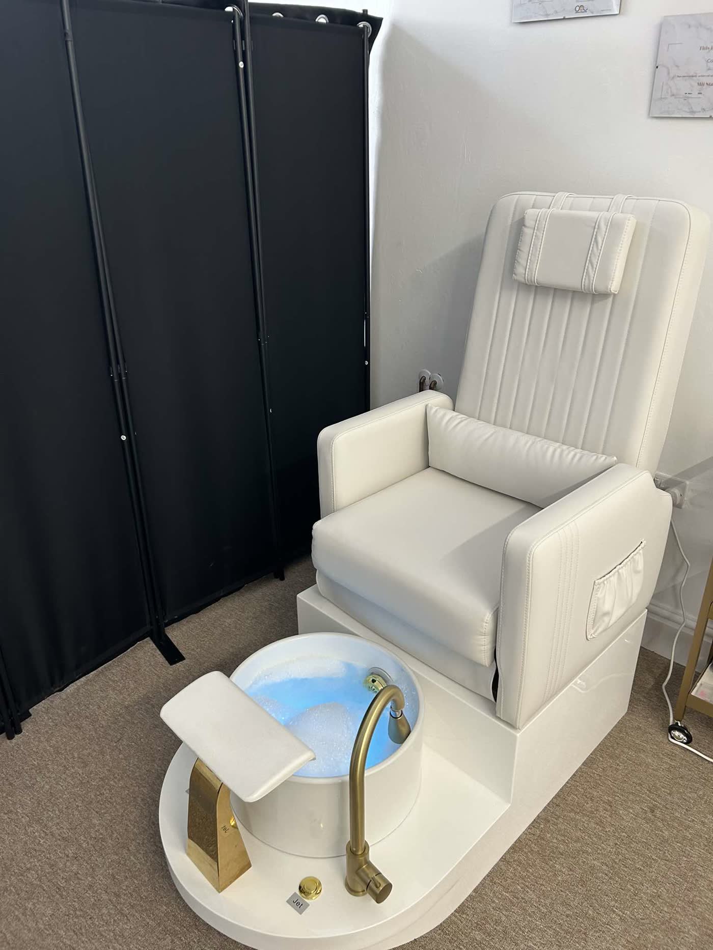 Pedicure spa station — white reclining chair with pillow and gold-tap foot bath, dark privacy screen behind.